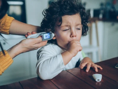 Małe dziecko siedzące przy stole w piżamce i kaszlące w piąstkę, w trakcie gdy mama sprawdza mu temperaturę