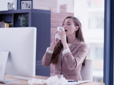 Kobieta siedząca w pracy przed komputerem, kichająca w chusteczkę
