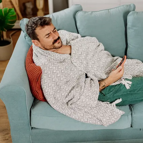 Ein mittelalter Mann sitzt in eine Decke gehüllt auf der Couch und putzt sich die Nase.